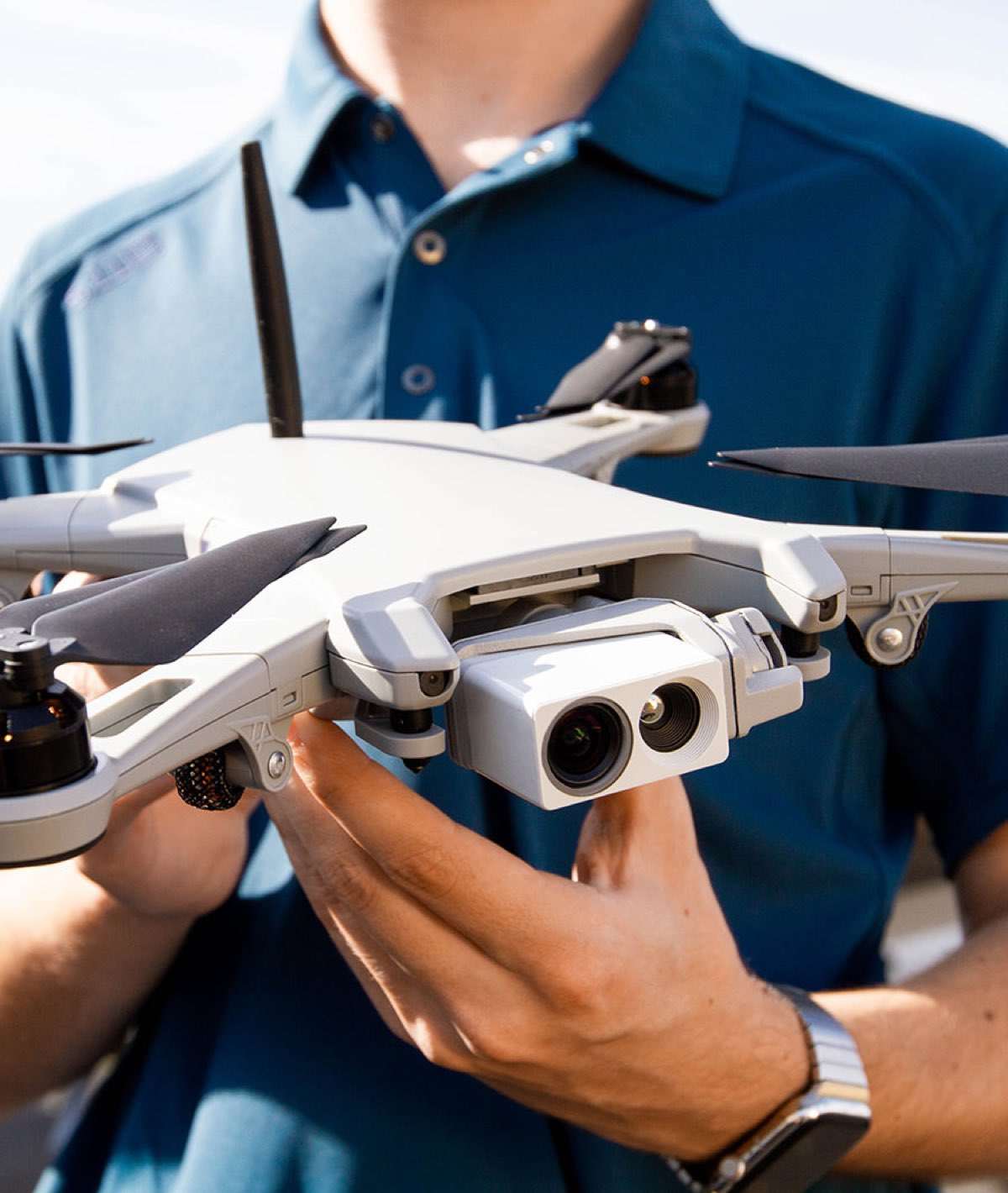 A person holding a multirotor aircraft in both hands, its arms extended and propeller blades folded back, with a dual-sensor camera payload at the front